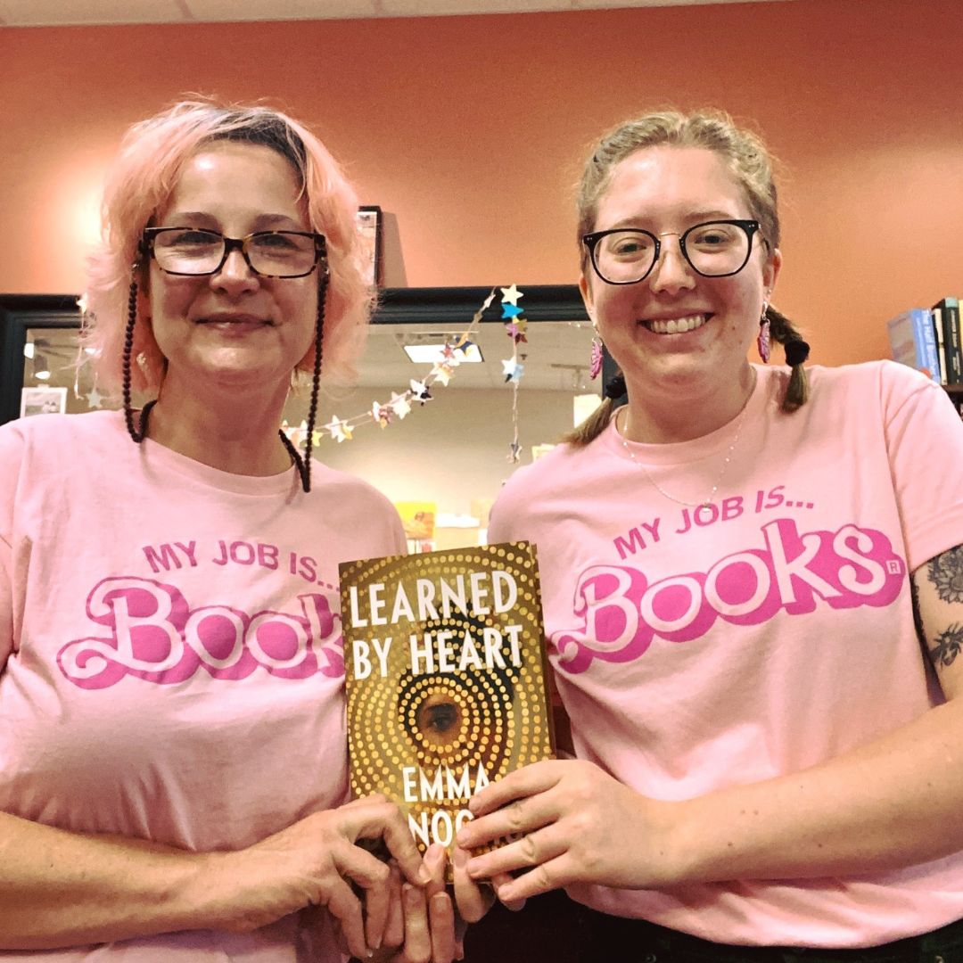 Store Manager Ellen Woodall and bookseller Molly Larson wearing pink shirts that say 'My job is Books' in Barbie style and holding up a copy of Learned by Heart by Emma Donoghue